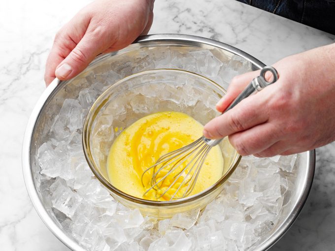 hands whisking eggs in a glass bowl that rests in a larger bowl filled with ice