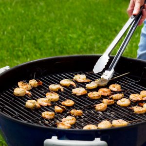 Person using metal tongs to flip skewered shrimp on a charcoal grill