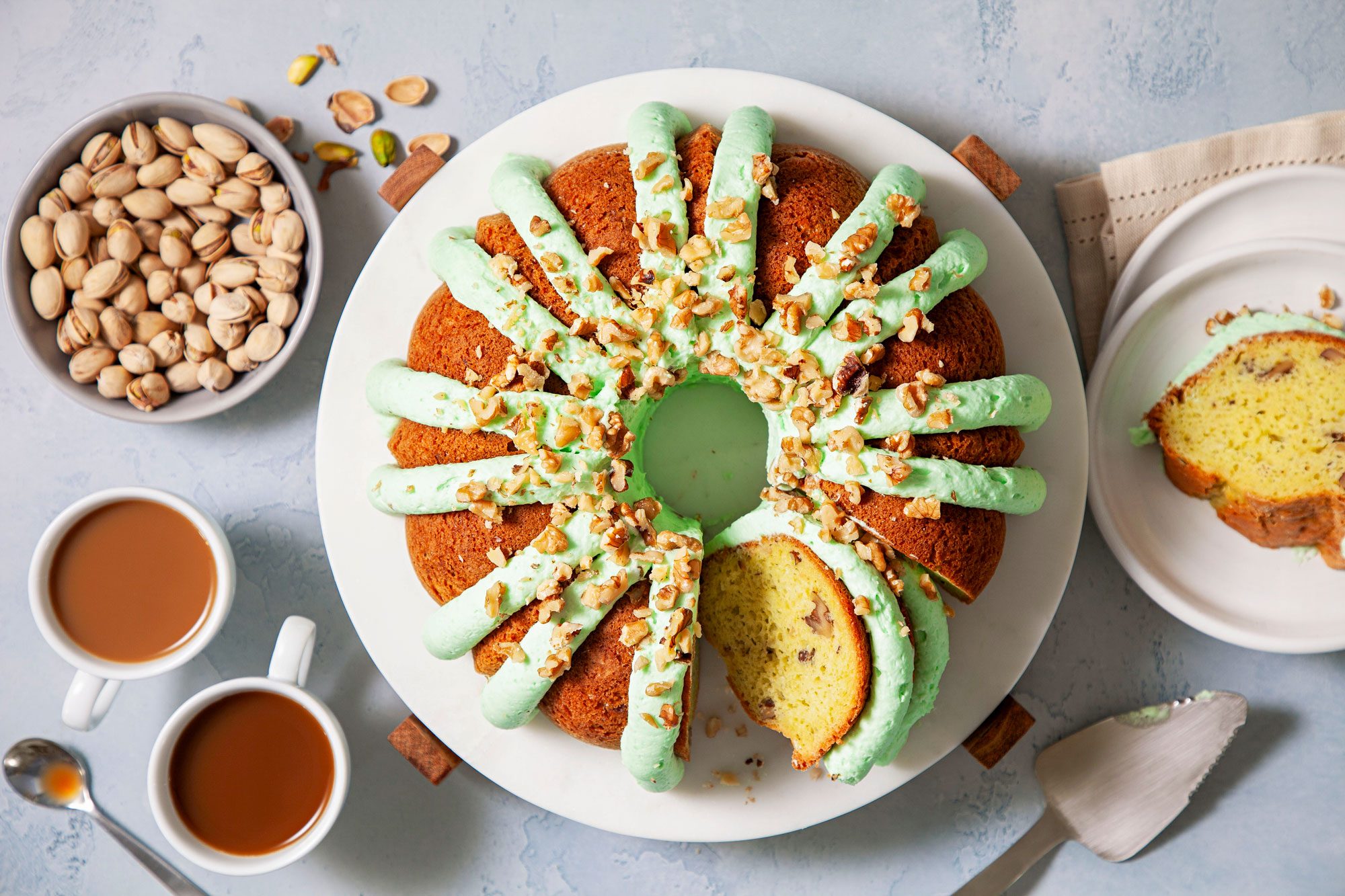 Pistachio Pudding Cake On A White Plate with A Bowl of Pistachio and Coffee Cups Next to it