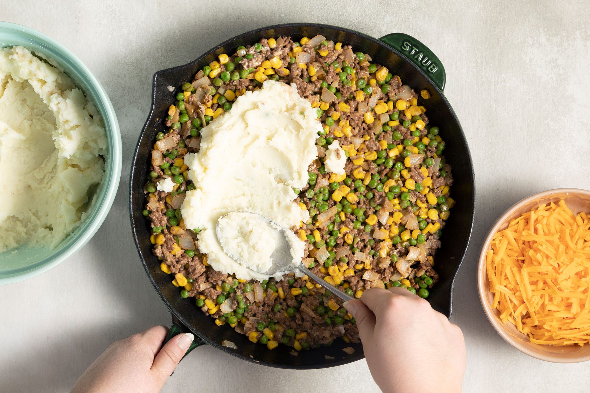 someone spreading the creamy mashed potatoes over the top of the mixture of cooked beef, corn, peas in a large skillet on induction cooktop
