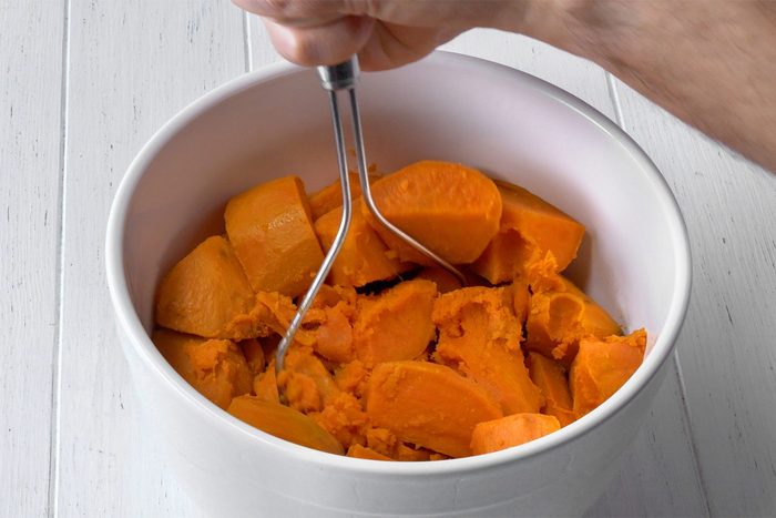 A close-up image of a person mashing chunks of cooked sweet potatoes in a white ceramic bowl using a stainless steel potato masher. The bowl is placed on a white wooden surface. The person’s hand gripping the masher is visible in the top right corner.