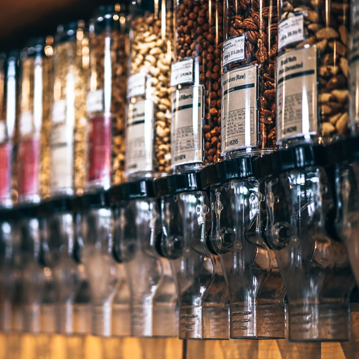 Glass jars full of different nuts and seeds are placed in row on shelf of a grocery store.