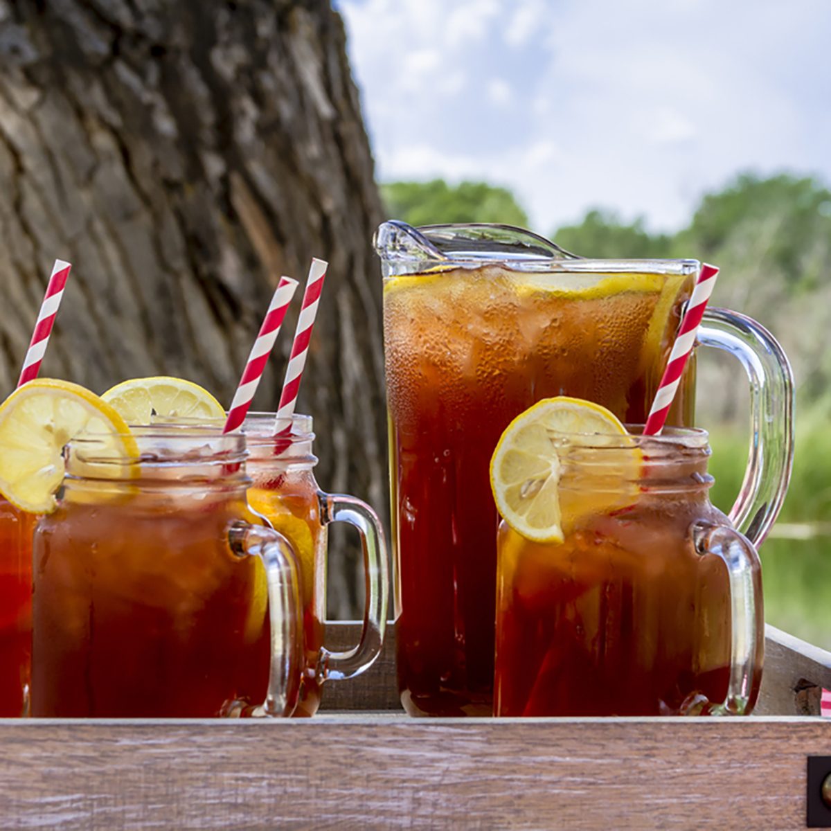 Pitcher and mason jar mugs filled with iced tea and lemons sitting on picnic table with red checked tablecloth
