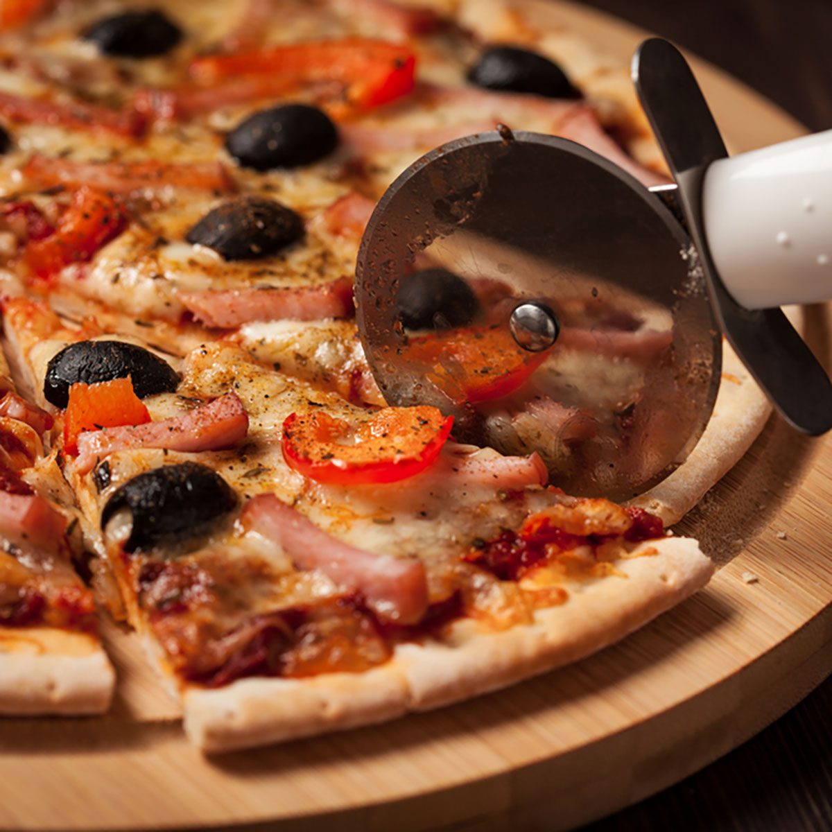 Pizza cutter (wheel) slicing ham pizza with capsicum and olives on wooden board on table; Shutterstock ID 249370456; Job (TFH, TOH, RD, BNB, CWM, CM): Taste of Home
