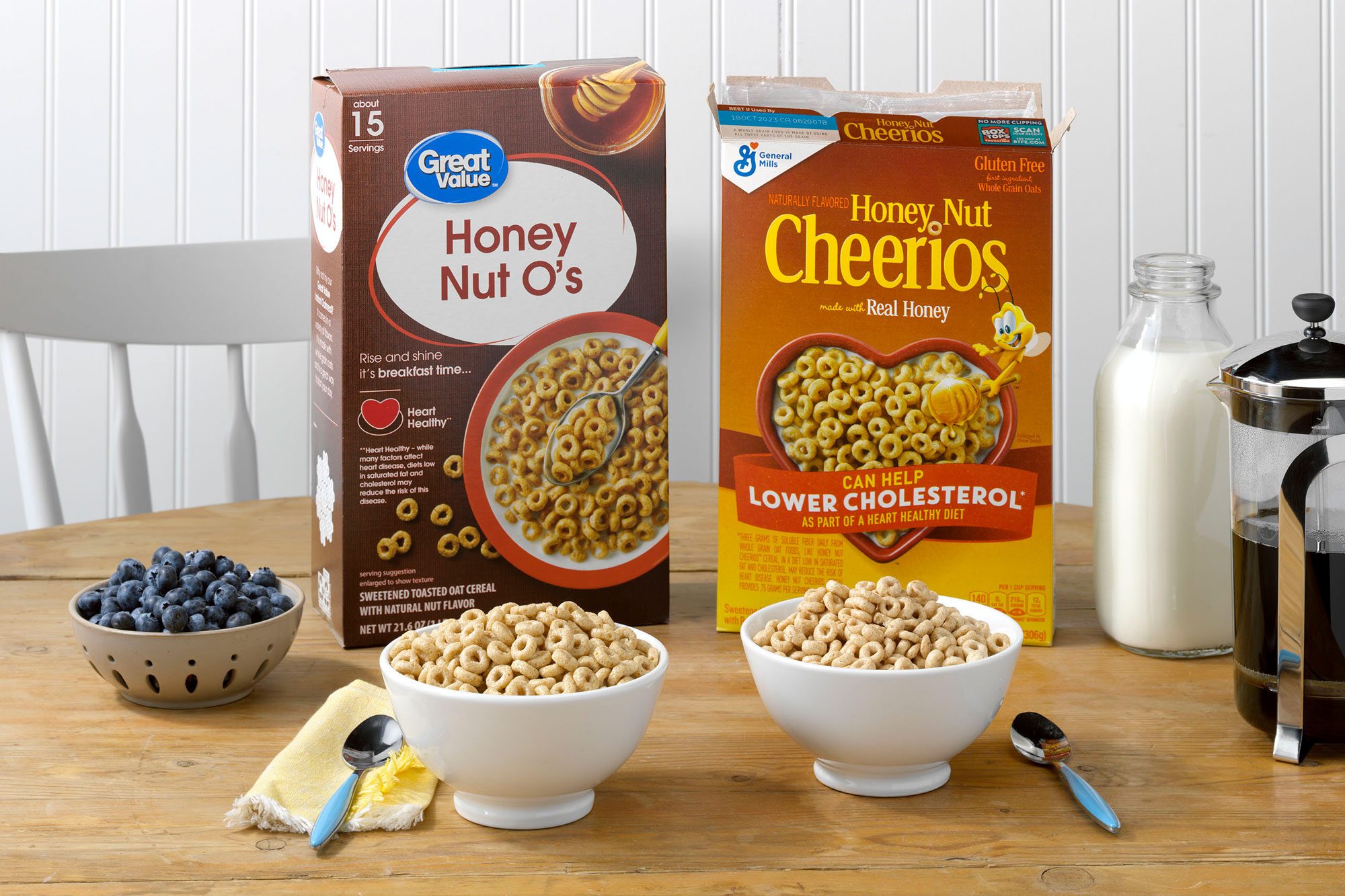 General Mills' Honey Nut Cheerios and Walmart's Great Value Honey Nut O's on a table with milk, coffee and blueberries