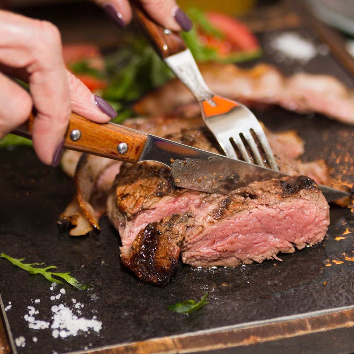 Woman hand holding knife and fork cutting grilled beef steak on stoned plate.