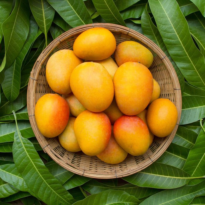 Mango tropical fruit in wooden basket put on green leaf background, top view