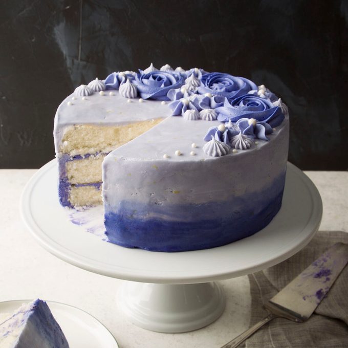 A frosted cake with shades of blue and purple on a white cake stand. The cake has a slice removed, revealing its interior layers. Flower and swirl decorations adorn the top, with a dark background behind.