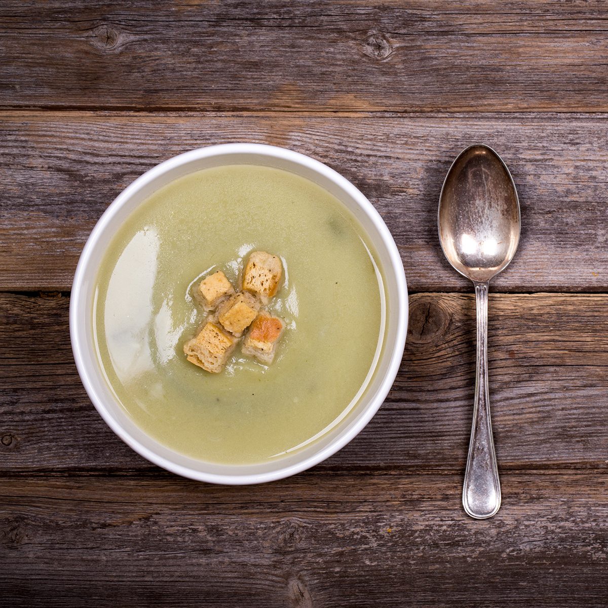 A bowl of leek and potato soup with bread croutons, over old wood table with fresh leeks and potatoes alongside