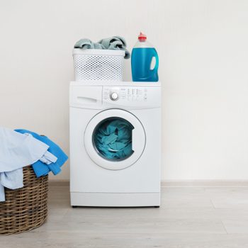 Photo of preparation to laundry process. White washing machine standing on laminate floor near wooden basket with clothes. Object isolated against wall inside bright light flat interior