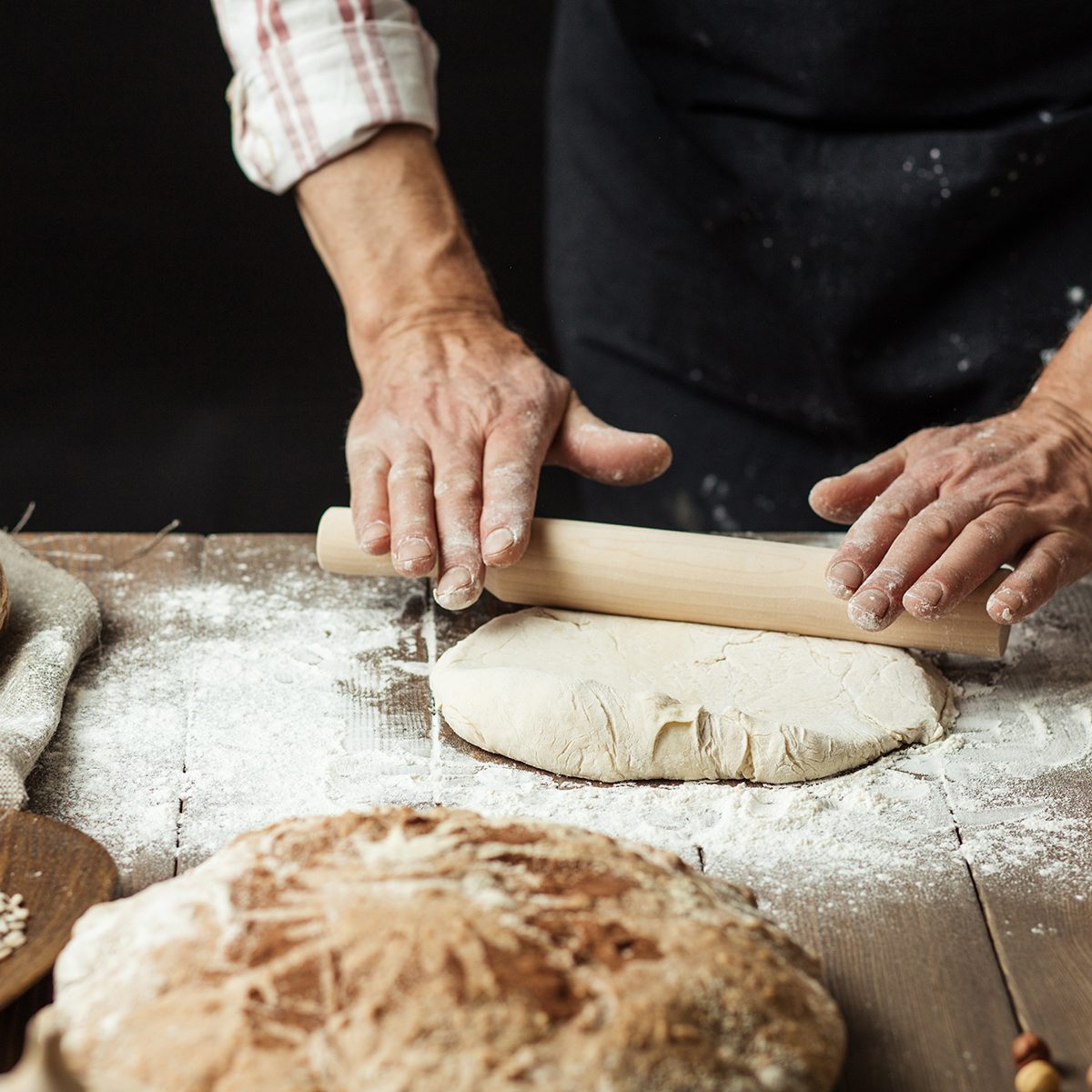 Chef or baker, dressed in black apron, preparing a portion of fresh dough in rural bakery, kneading the pastry surrounded by rustic organic loaf of bread.