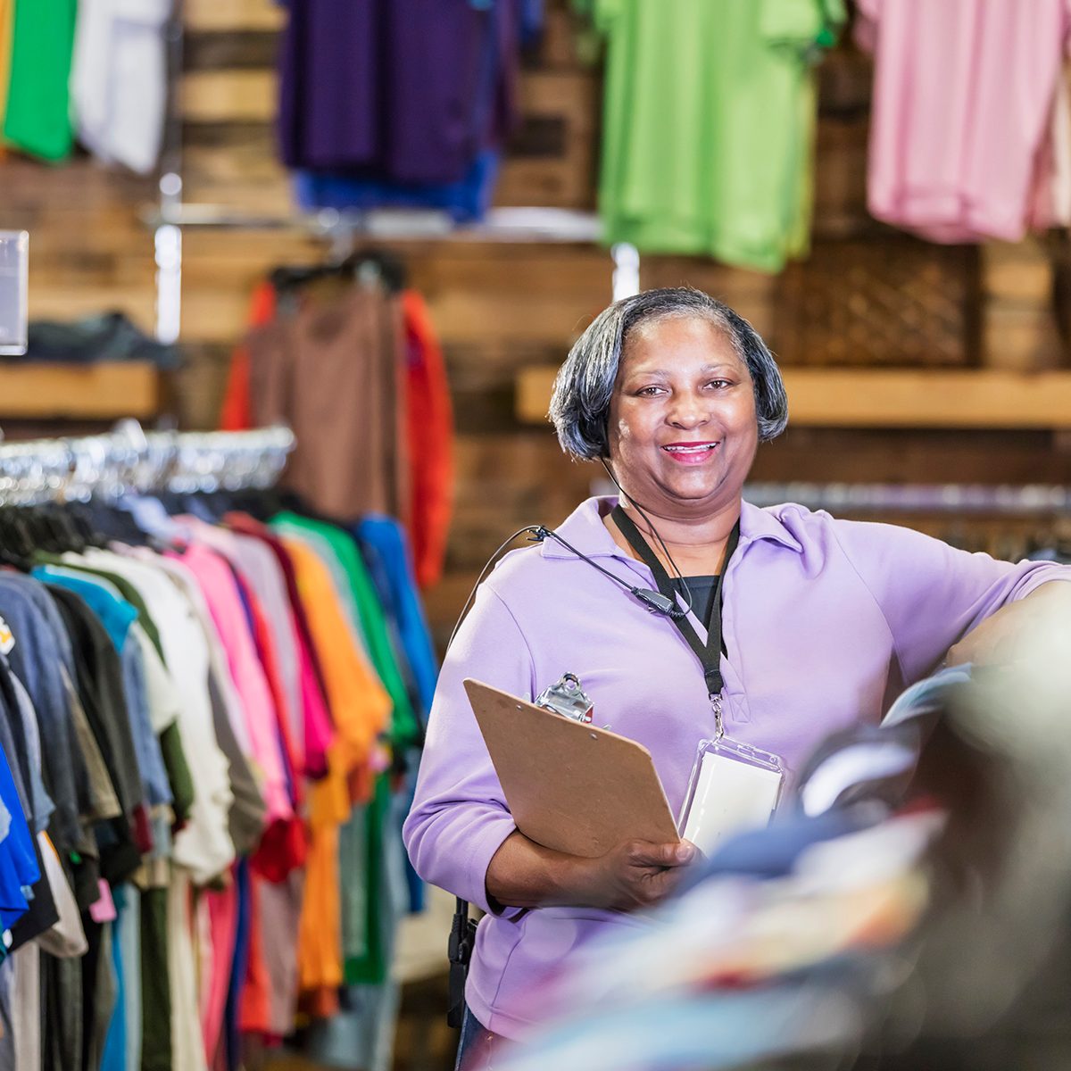 A senior African-American woman in her 60s working in a clothing store. She is standing in the menswear department at a rack of clothing, holding a clipboard.