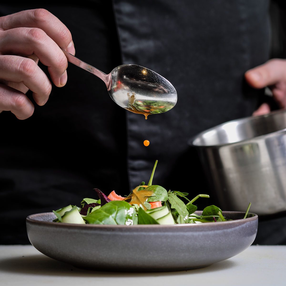 Close-up of the hands of a male chef on a black background. Pour sauce from the spoon on the salad dish. Food decoration.