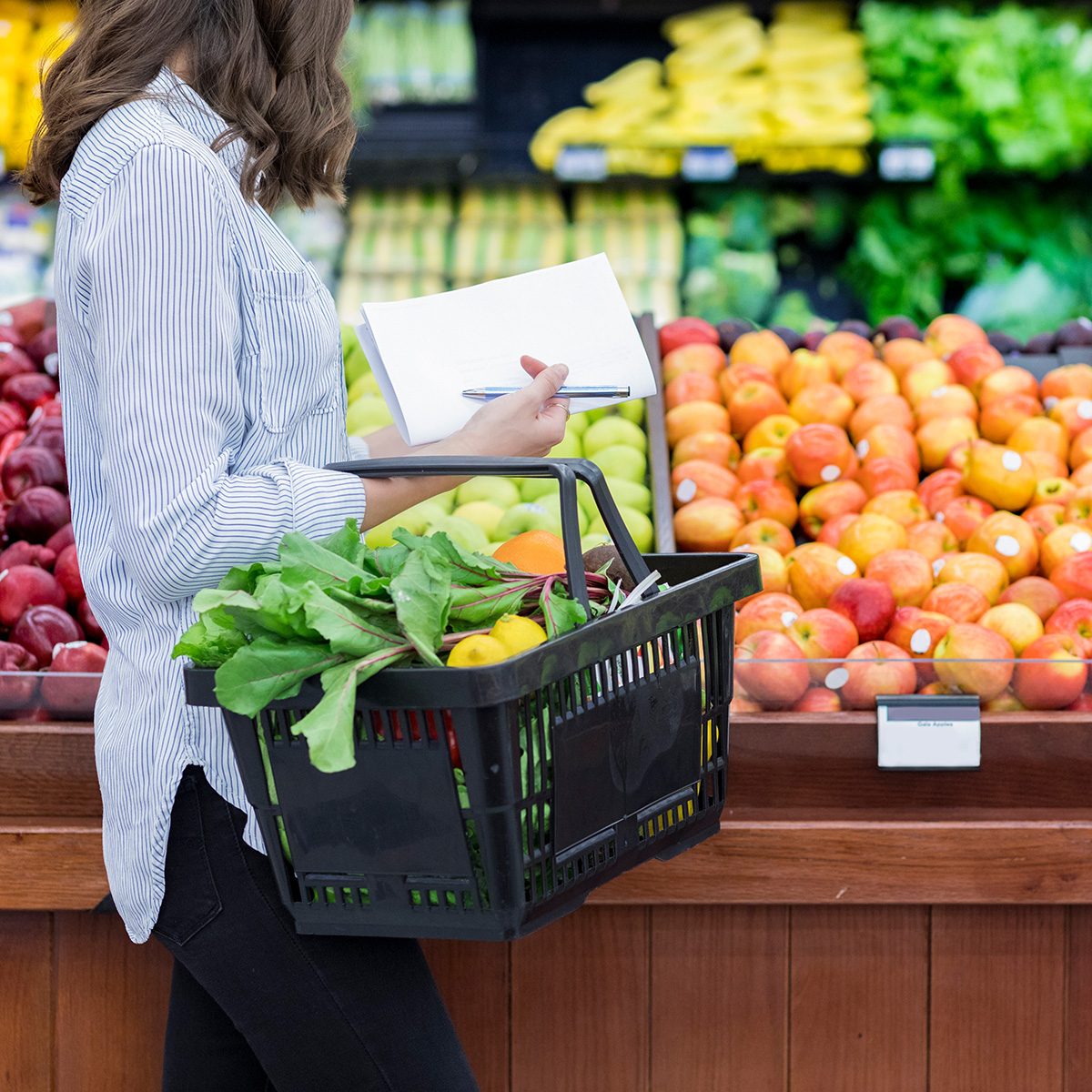 Young woman carries a shopping basket filled with fresh produce. She is shopping for fresh fruit and vegetables in a grocery store.