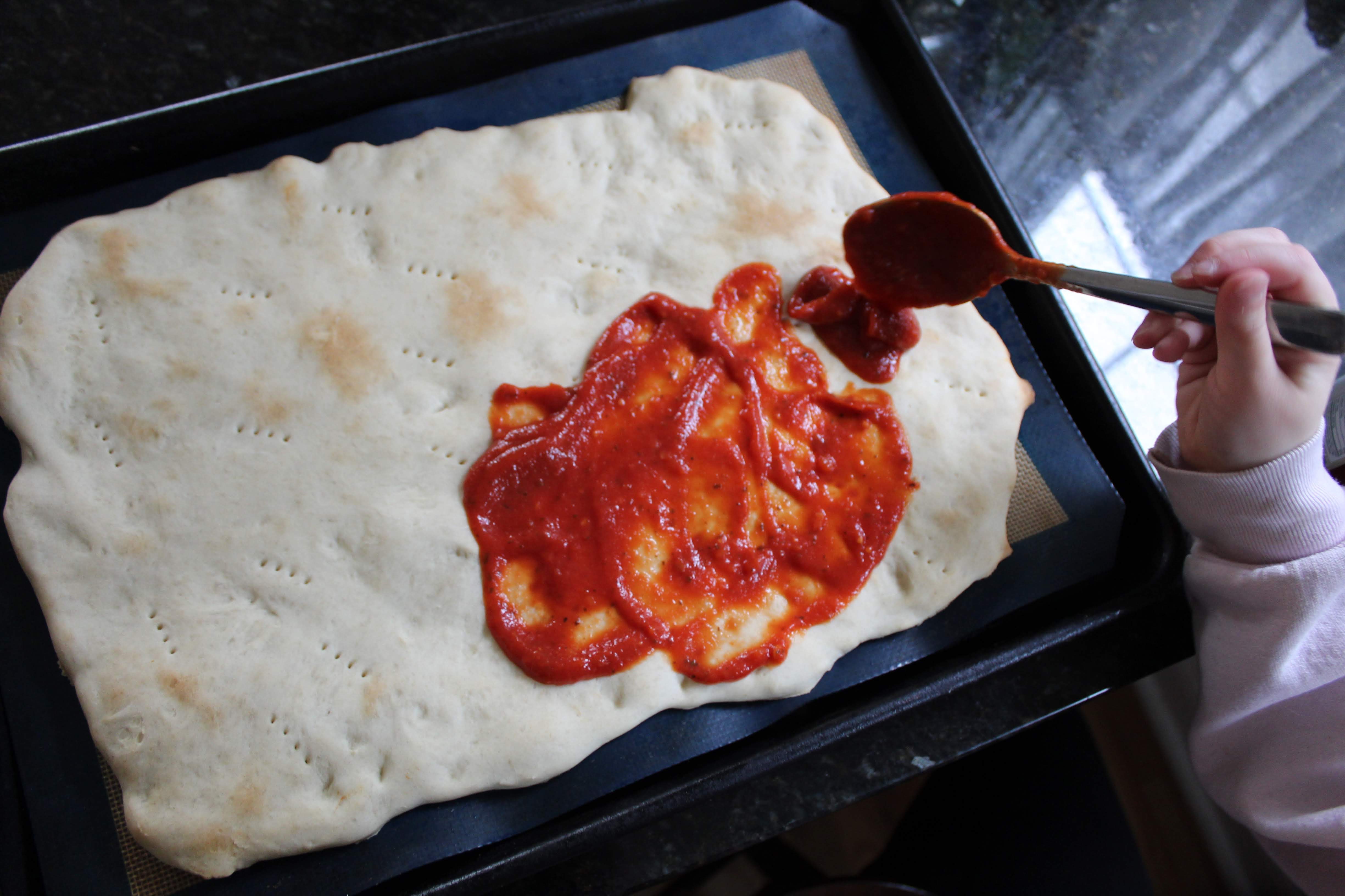 Child hands spreading pizza sauce on crust with a spoon