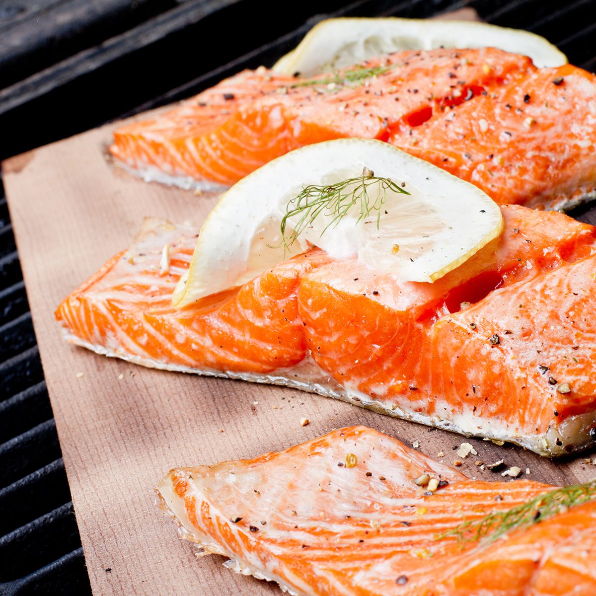 Three wild-caught salmon filets on a cedar plank in a backyard grill. Fist is bright reddish orange and is topped with lemon, dill and cracked pepper