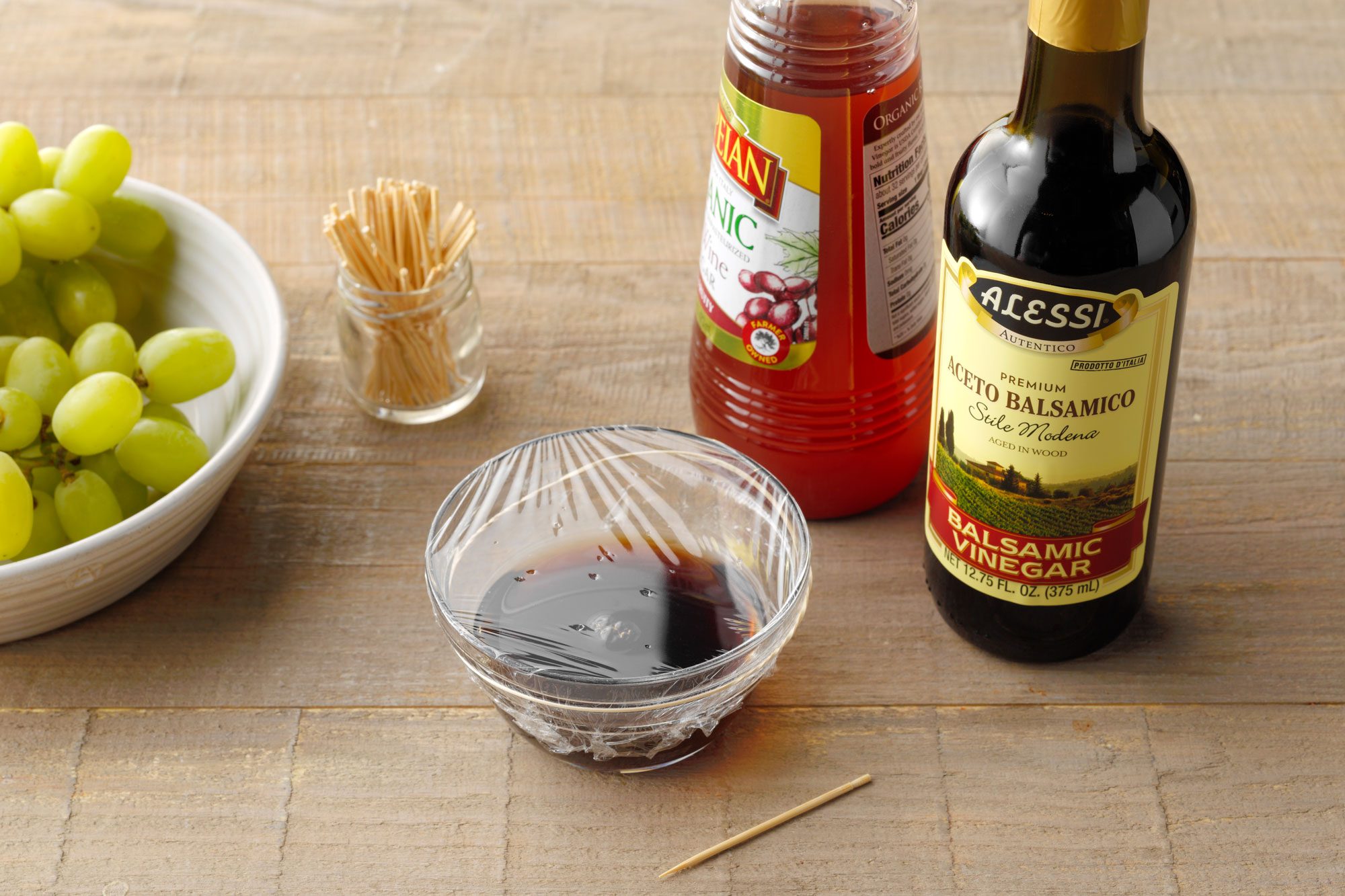 trap 3 mixture in a small glass bowl covered with plastic wrap that has holes poked into it. fresh grapes, toothpicks, red wine vinegar bottle, and balsamic vinegar bottle nearby. all on a wooden surface