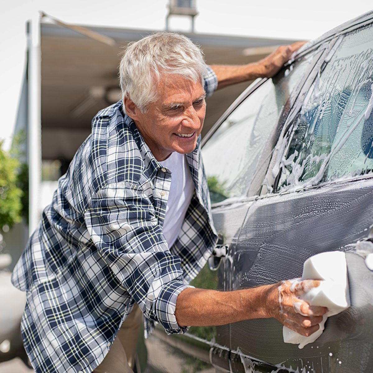 Happy senior man washing car with soap and foam. Old retired man cleaning automobile with sponge in a sunny day. Satisfied driver washing his gray car near the garage.
