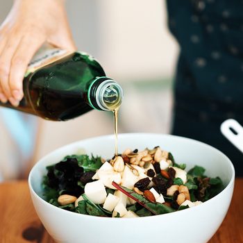 how to store olive oil Close-up view of a bowl of fresh green salad with mozzarella, mixed nuts and dry fruits. A woman’s hand was pouring olive oil into the salad.