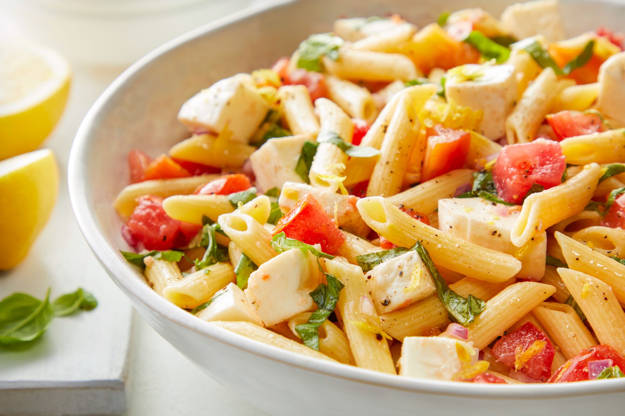 Caprese Pasta Salad in a Large Ceramic Bowl with Sliced Lemon and Basil on Left