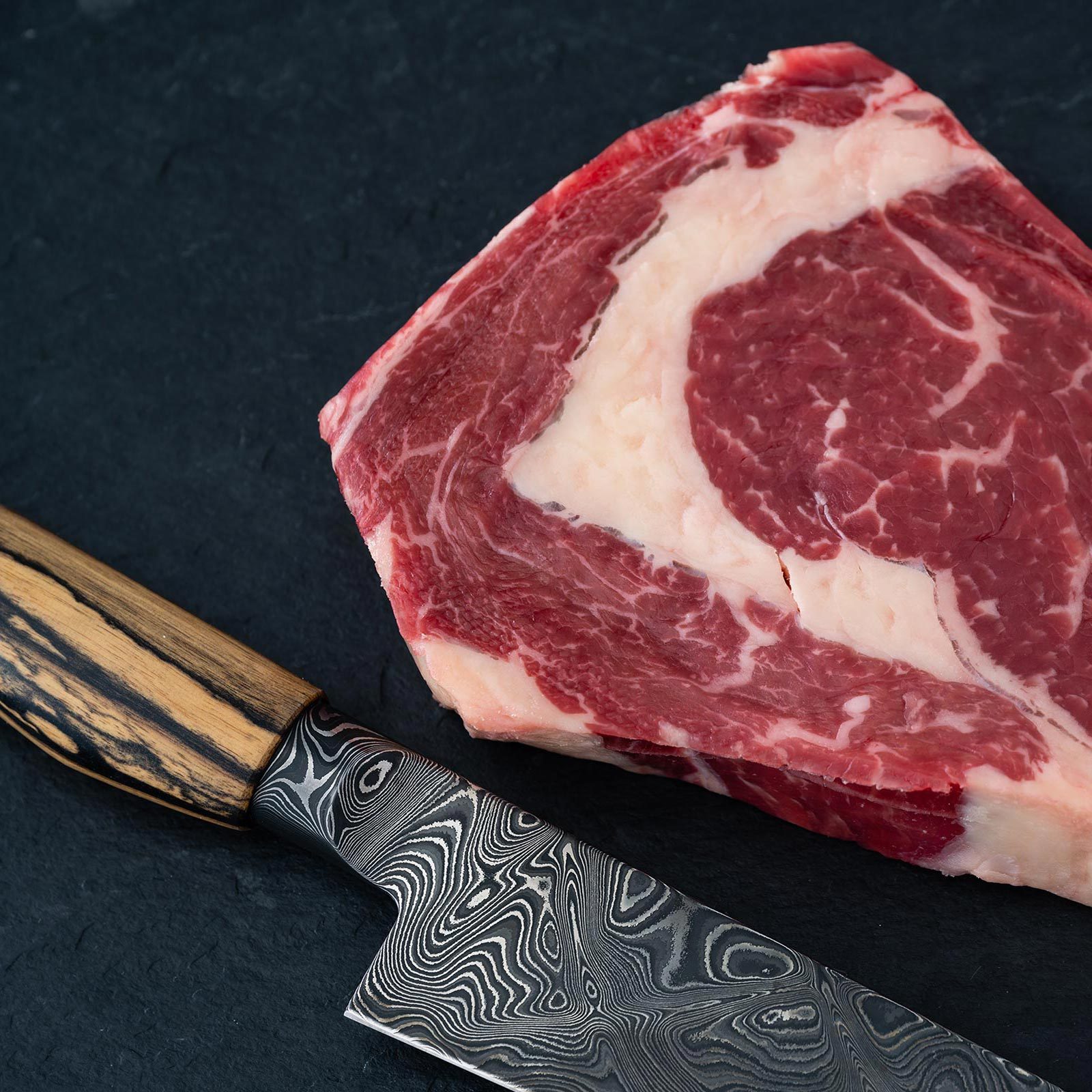 Raw Wagyu Beef Ribeye Steak on a Dark background with a damascus kitchen knife