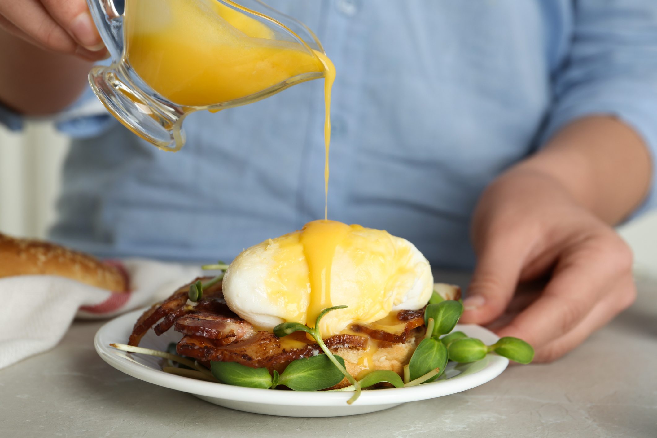 Woman pouring Hollandaise sauce onto egg Benedict at table, closeup