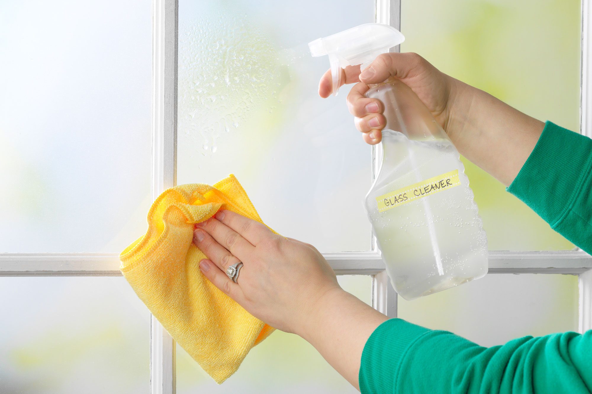 hand using homemade glass and windows cleaner to spray windows at home. holding yellow microfiber cloth in other hand