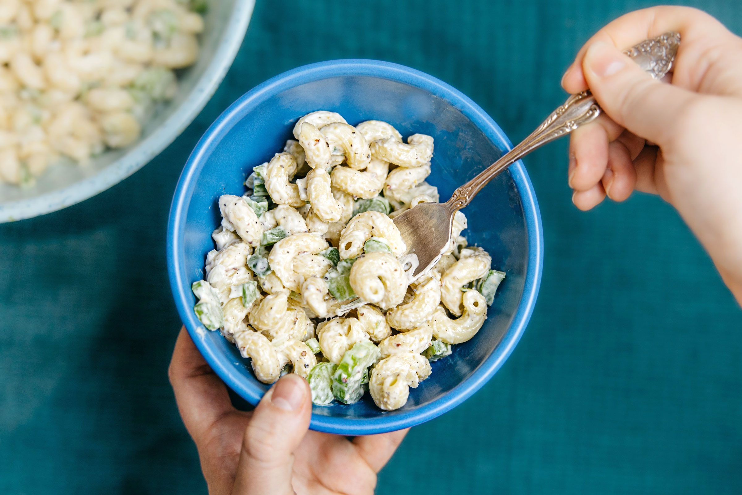 hand with wooden spoon with Mac salad in a blue bowl on a matching blue towel with a larger bowl of Mac salad of to the side