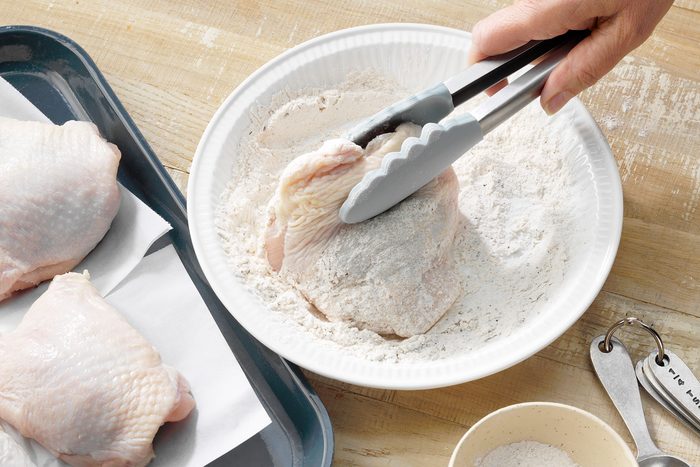 A raw chicken thigh being coated in flour in a white bowl using metal tongs. To the left, additional raw chicken thighs rest on a parchment-lined tray. Measuring spoons and a small bowl of flour are placed on the wooden surface nearby.