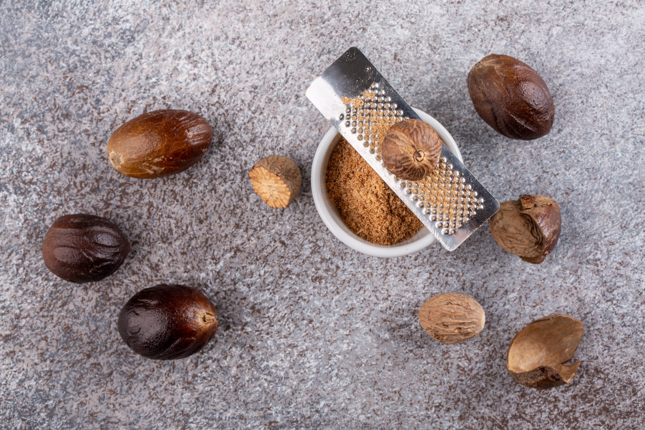 Nutmeg seed and ground nutmeg with grater on concrete background. Freshly grated spice in white bowl. Close-up, top view.