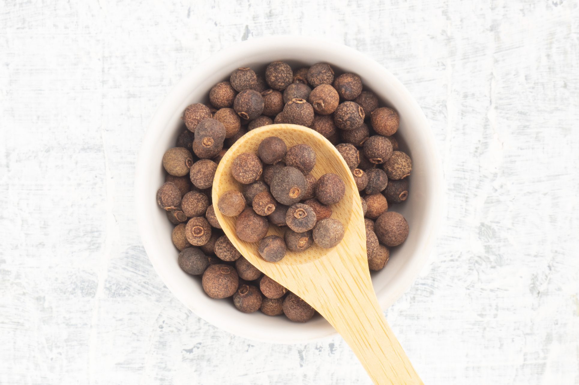 Spice Allspice (Jamaica pepper, Pimento) in ceramic bowl and wooden spoon on white concrete background. Top view. Indian cuisine concept