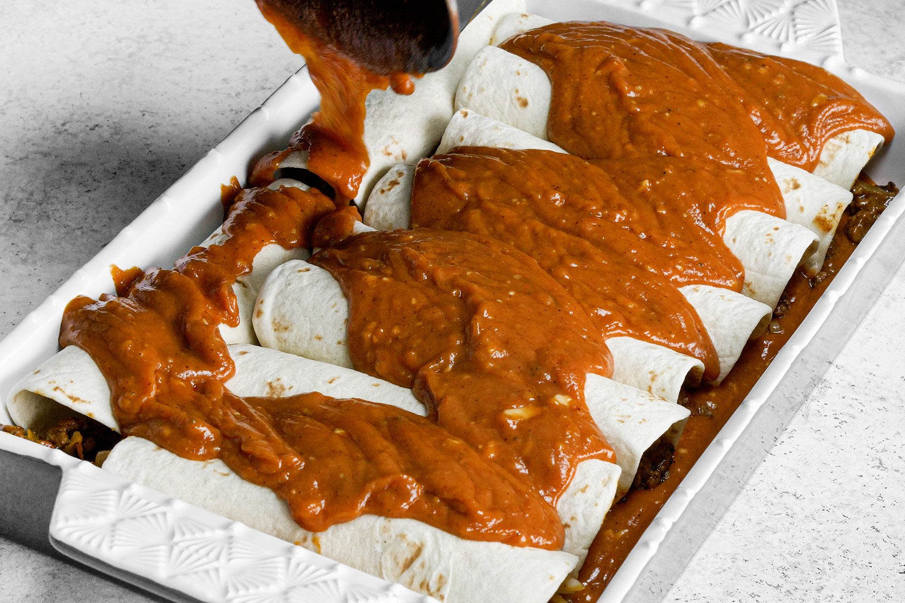 Pouring sauce on the rolled up tortillas on baking dish