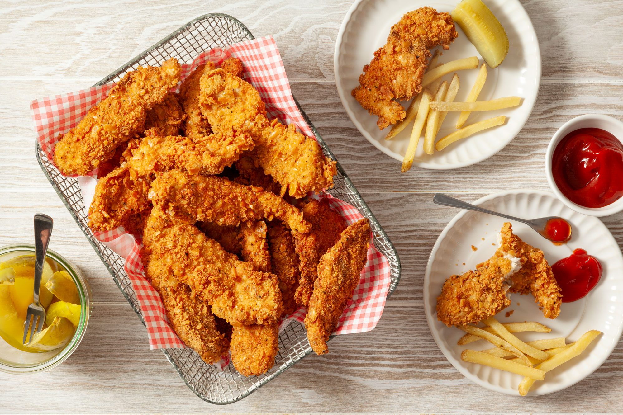 Fried Chicken Strips in Wire Mesh Basket on a Wooden Surface and Fried Chicken Strips in Two Small Plates with French Fries and Tomato Ketchup Next to it.