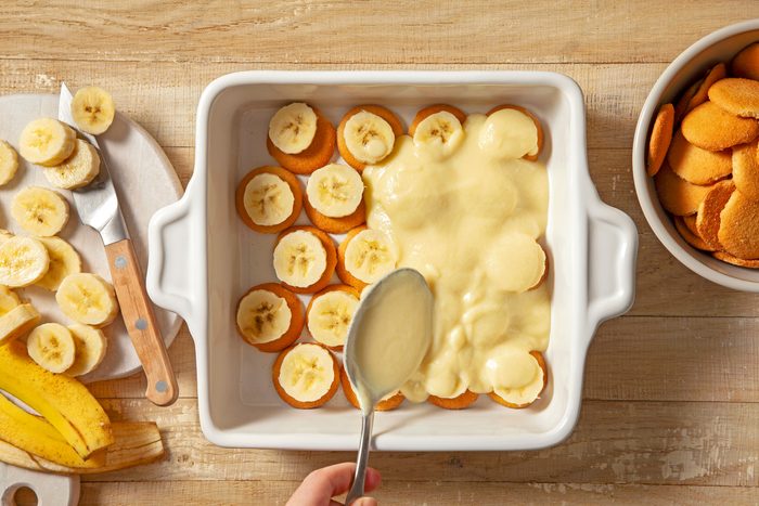 A hand is layering pudding over vanilla wafers and banana slices with a spoon in a baking dish