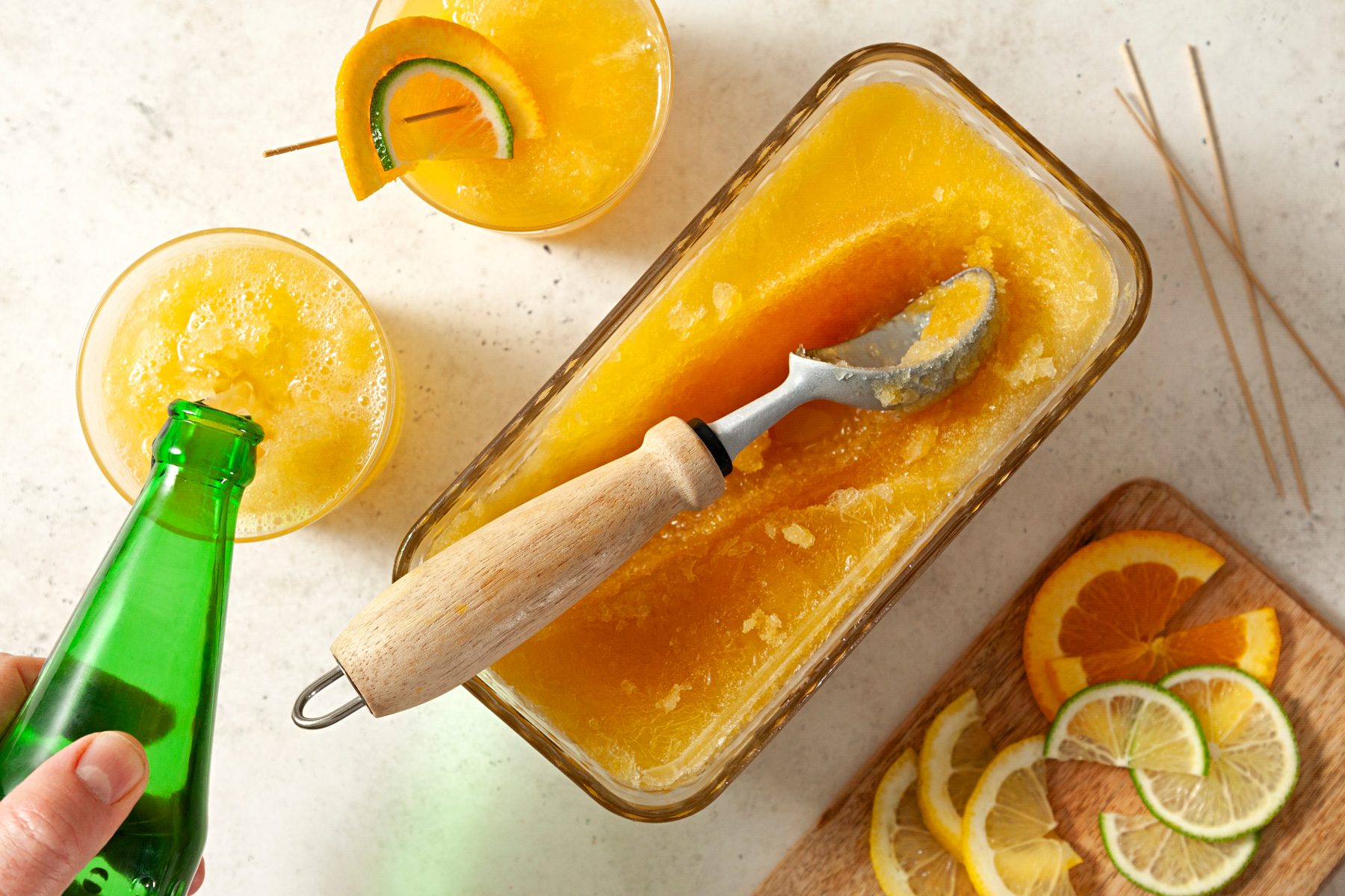 Freezed slush in container with slush served in glasses with orange and lemon wedges on wooden board.