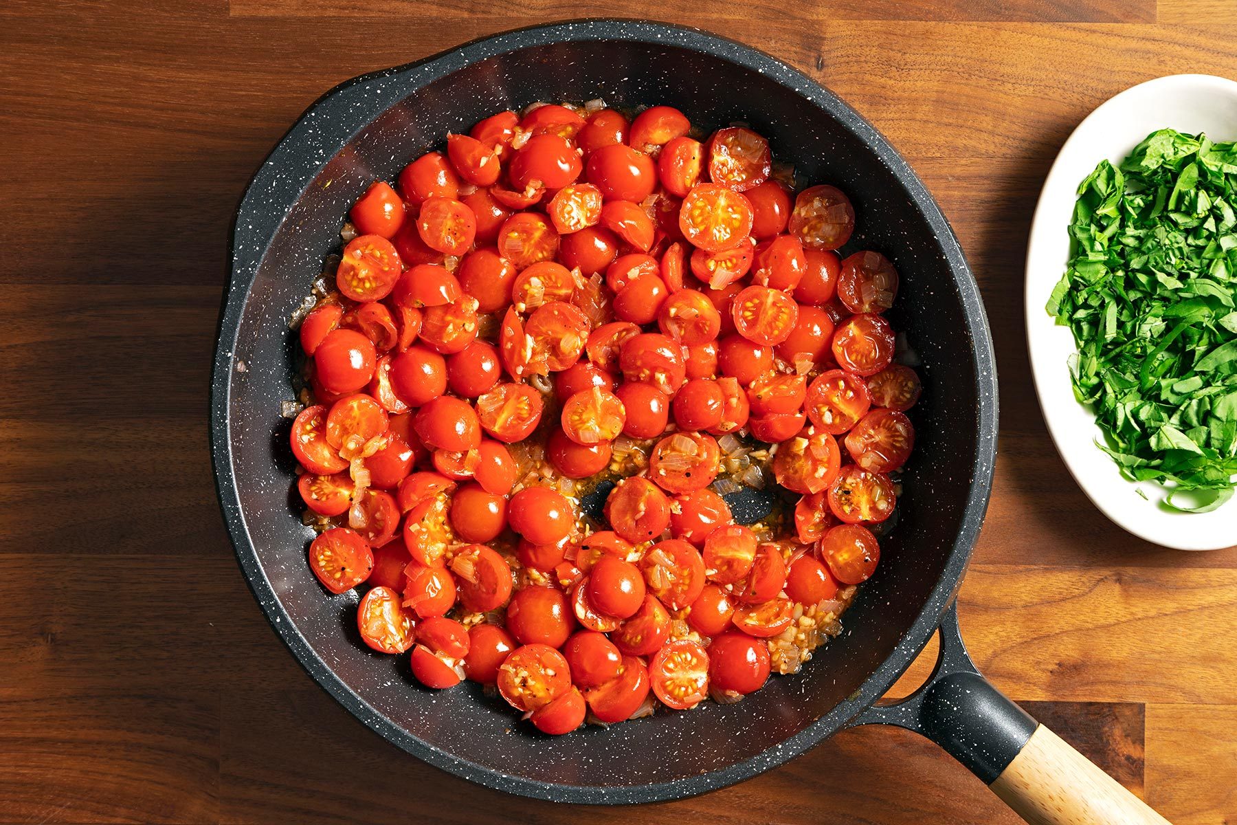 Overhead shot of topping in a large skillet; cook and stir unitl tomatoes are tender; bowl of chopped basil; brown wooden background;