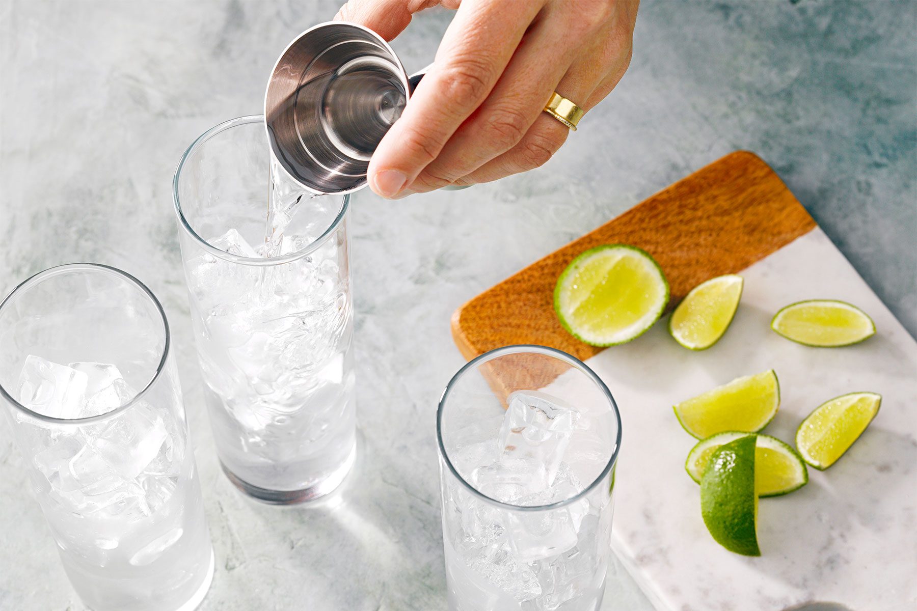 Overhead shot of fill highball or collins glasses with ice; pouring gin; lime wedges over chopping board; marble background;