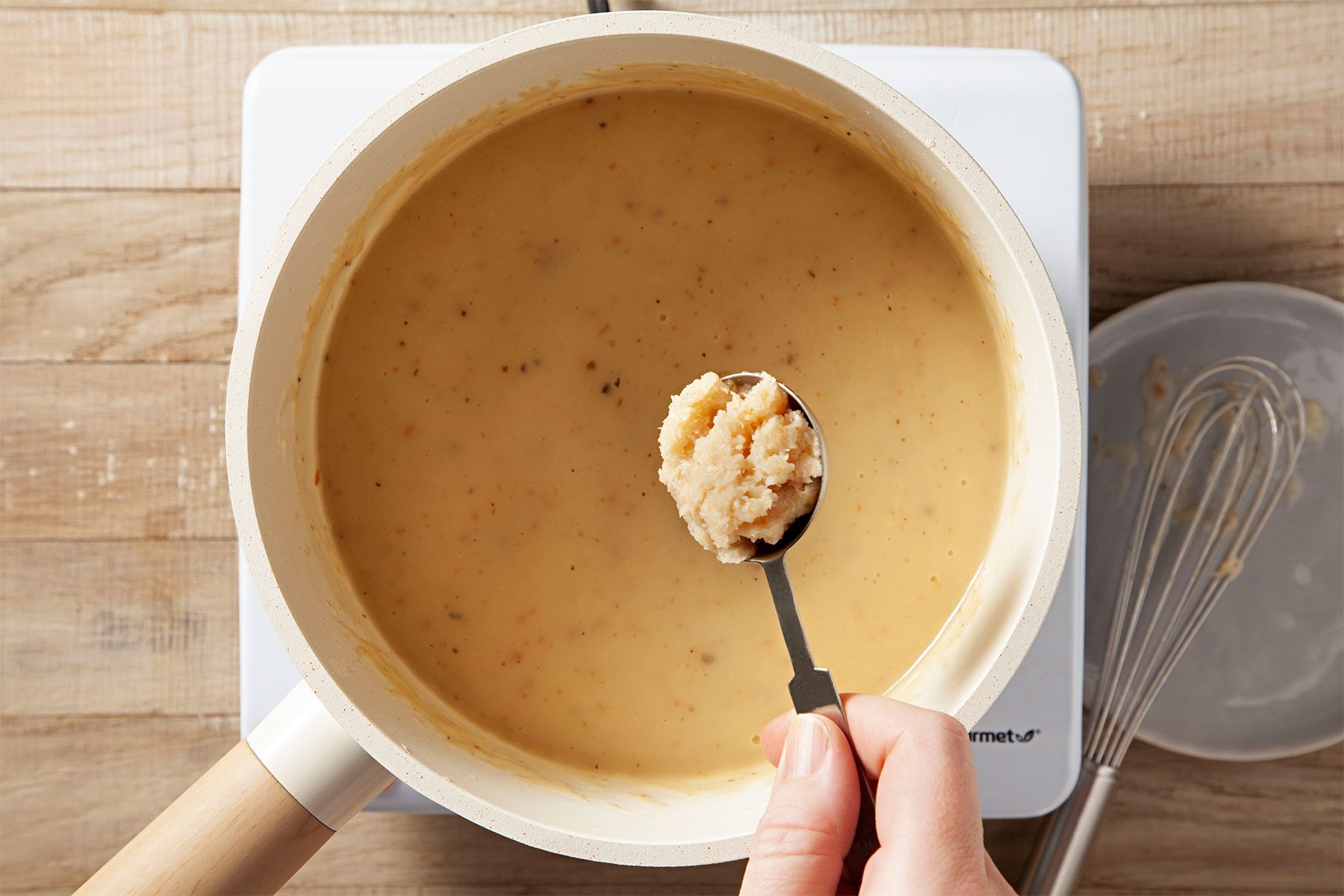 Overhead shot of a small saucepan; ham gravy; induction stove; whisk tool; wooden background;