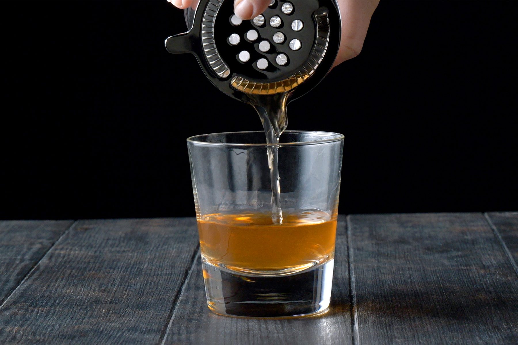 A hand holding a cocktail shaker with a strainer pours a light brown liquid into a clear glass filled halfway, set on a dark wooden table against a black background.