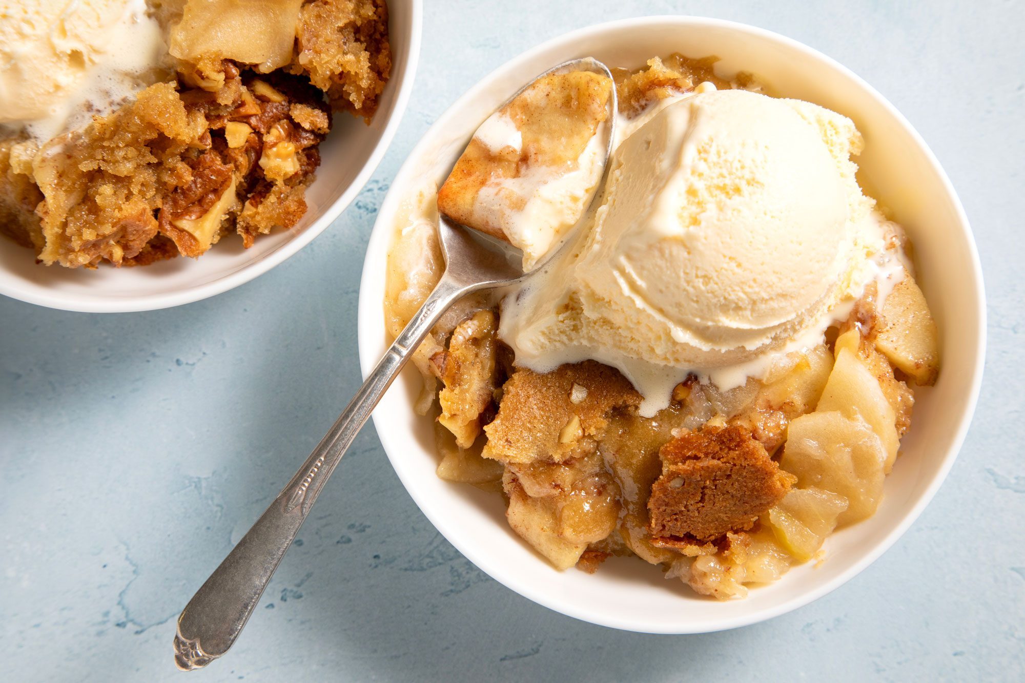 overhead shot; light blue textured background; Walnut Apple Dessert in two white bowls served with ice cream scoop with silver spoon;