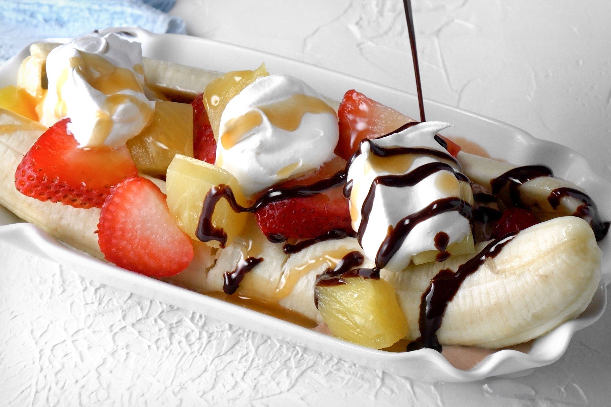 3/4th shot; white background; added strawberries, bananas and other ingredients, pouring chocolate syrup over the ice cream;