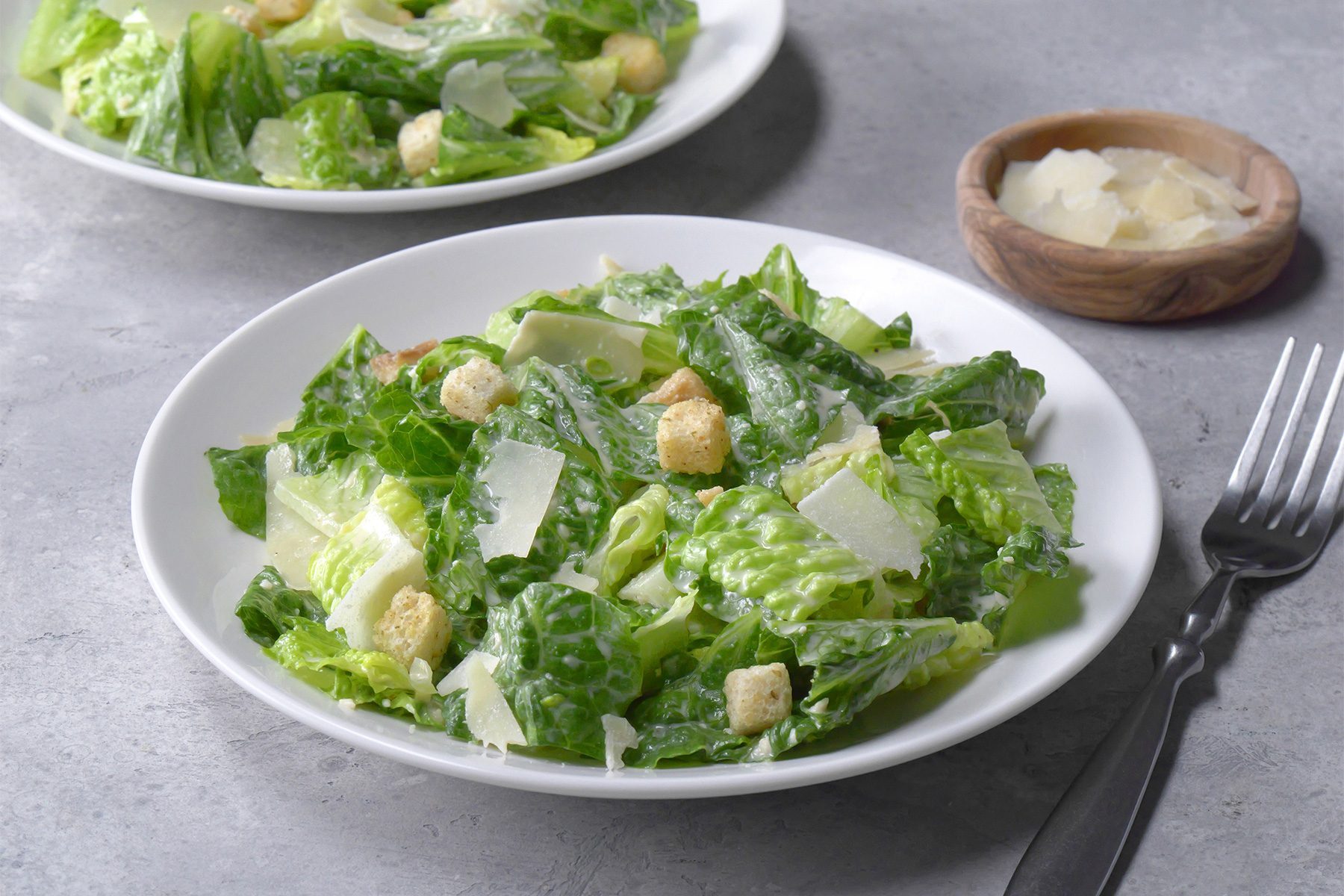 Overhead shot of Ceasar Salad; served on two white plates; sprinkle with croutons and parmesan; wooden spoon; white plate; fork; napkin; grey background
