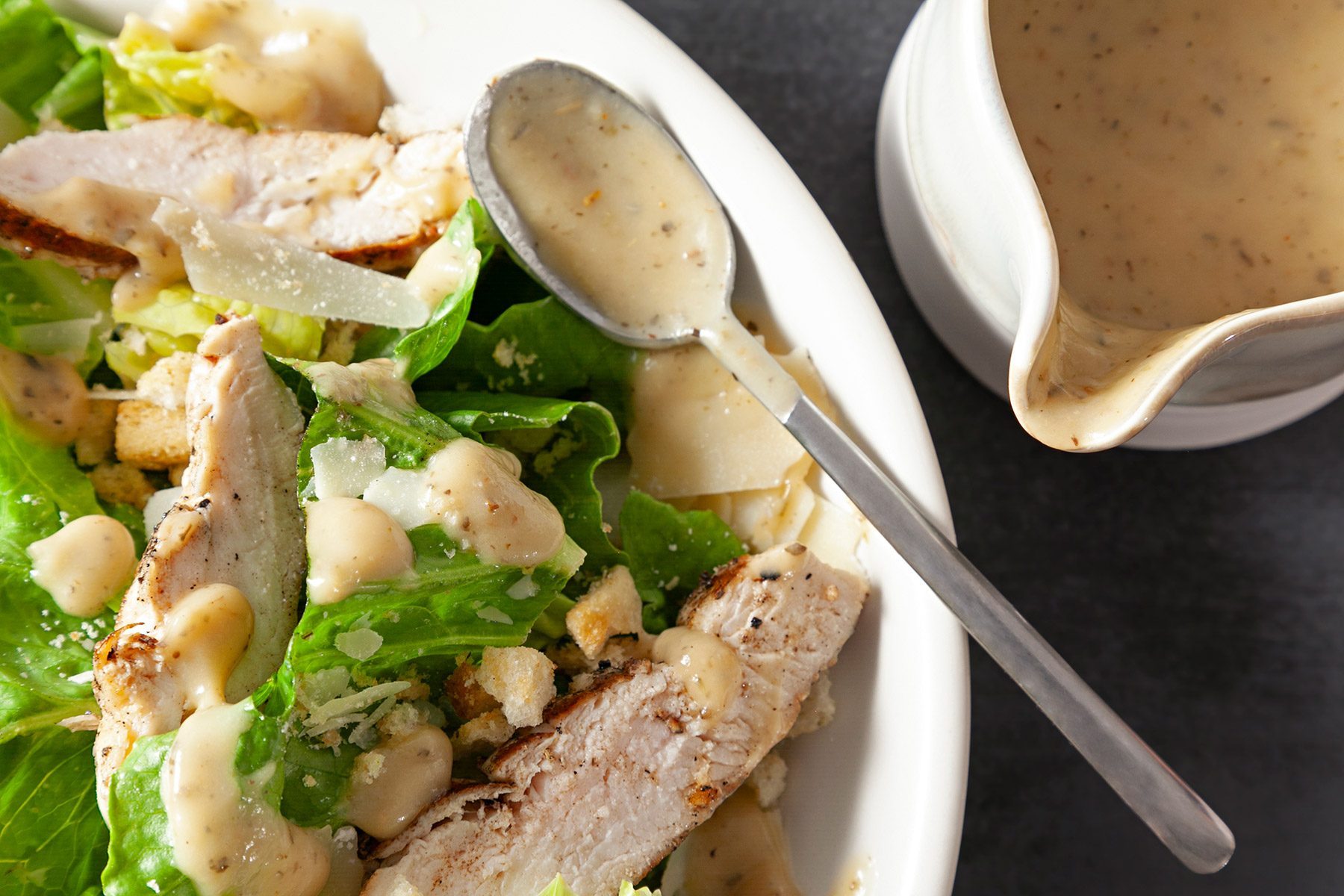 Top view shot of Roasted Garlic Vinaigrette; in a ceramic jar; dressing over salad; spoon; dark grey background
