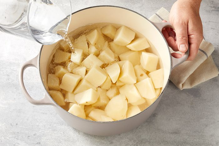 A pot filled with peeled and chopped potatoes placed on a countertop. A person's hand holds the pot handle while water is being poured into the pot, submerging the potato pieces. A cloth lies next to the pot.