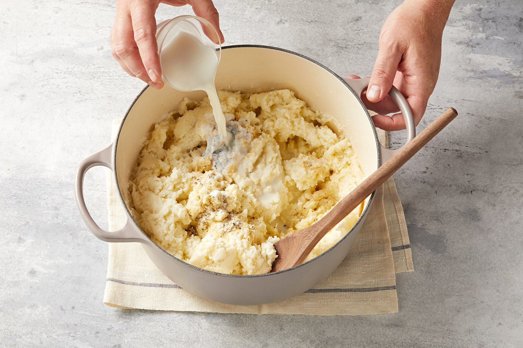 A person is pouring milk into a large pot of mashed potatoes. The pot is on a beige towel on a textured gray surface. A wooden spoon rests inside the pot.