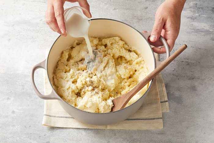A person is pouring milk into a large pot of mashed potatoes. The pot is on a beige towel on a textured gray surface. A wooden spoon rests inside the pot.