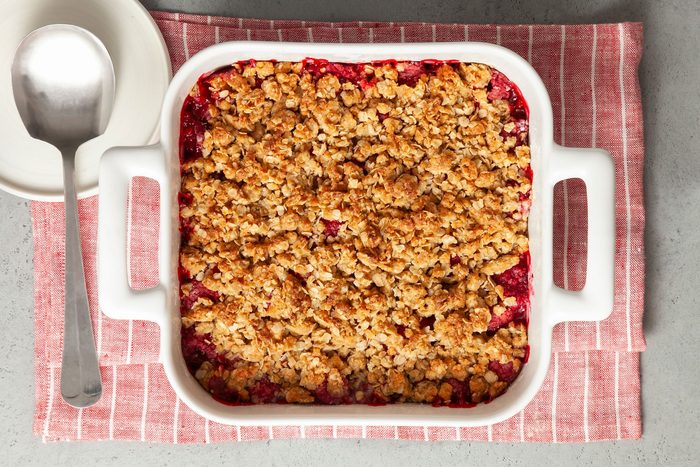 overhead shot; white background; Raspberry Crisp in a square baking dish over kitchen cloth with small white plate and a large serving spoon;