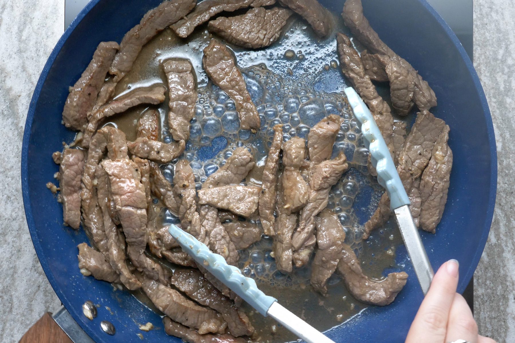A person stirring the steak strips with the ginger, garlic, and black pepper in a blue skillet