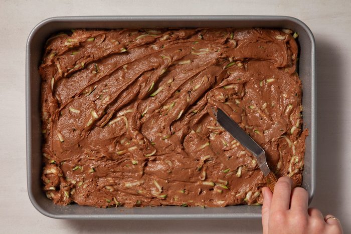 Overhead shot of pour Zucchini Brownies mix into a greased baking pan; cake knife; marble background;