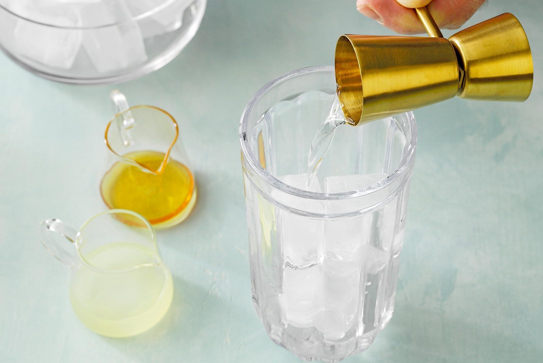 A hand holding a golden jigger is pouring a clear liquid into a tall glass filled with ice cubes. Nearby, two small pitchers containing yellow and green liquids are placed on a light blue surface.
